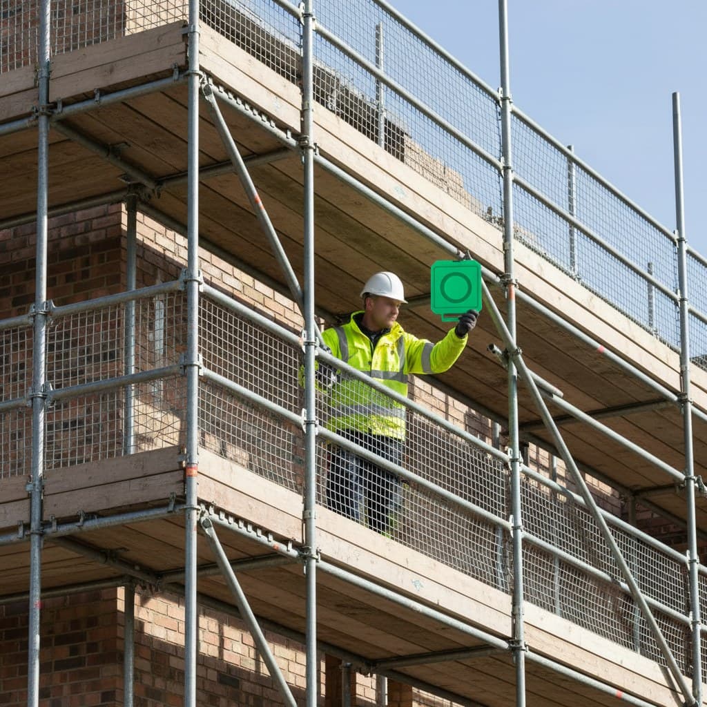 A scaffolder in a white hard hat and hi-vis jacket holding up a green scaffold tag on a fully guarded tube-and-fitting scaffold on a UK housing site