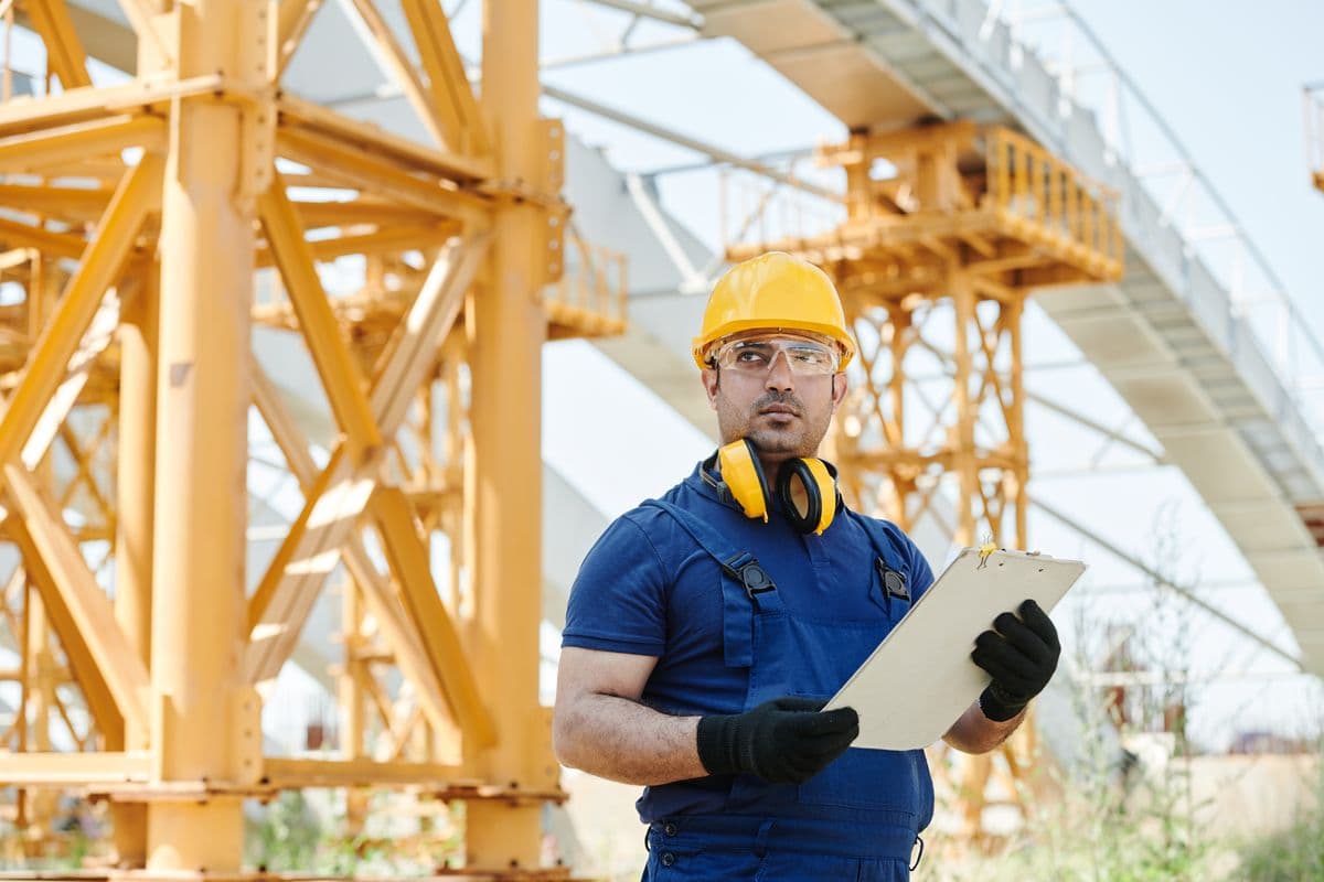 A worker in blue overalls, a yellow hard hat, safety glasses and ear defenders holds a clipboard in front of yellow steel formwork on a construction site.