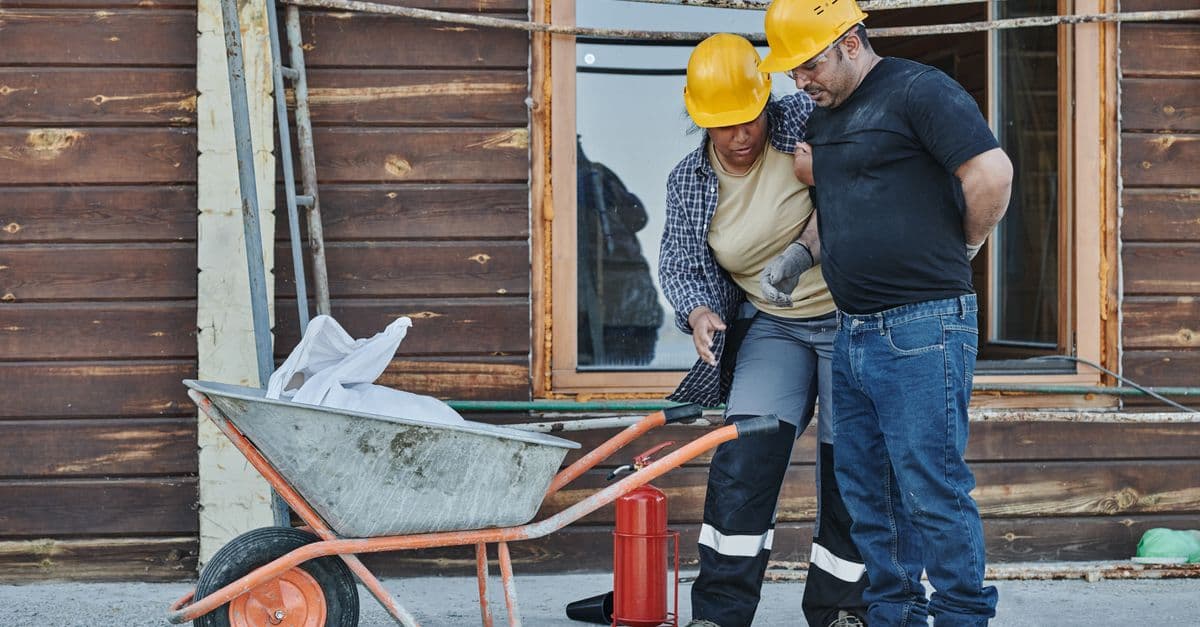 Two construction workers in hard hats helping an injured colleague, showing first aid support on site