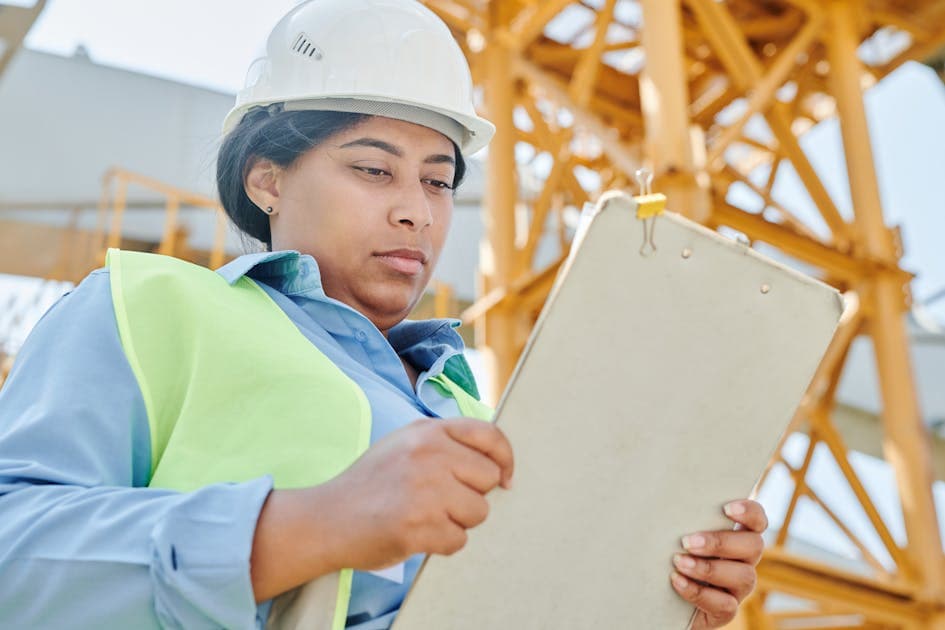 a confident female construction worker with clipboard and hard hat reviewing site plans on a construction site