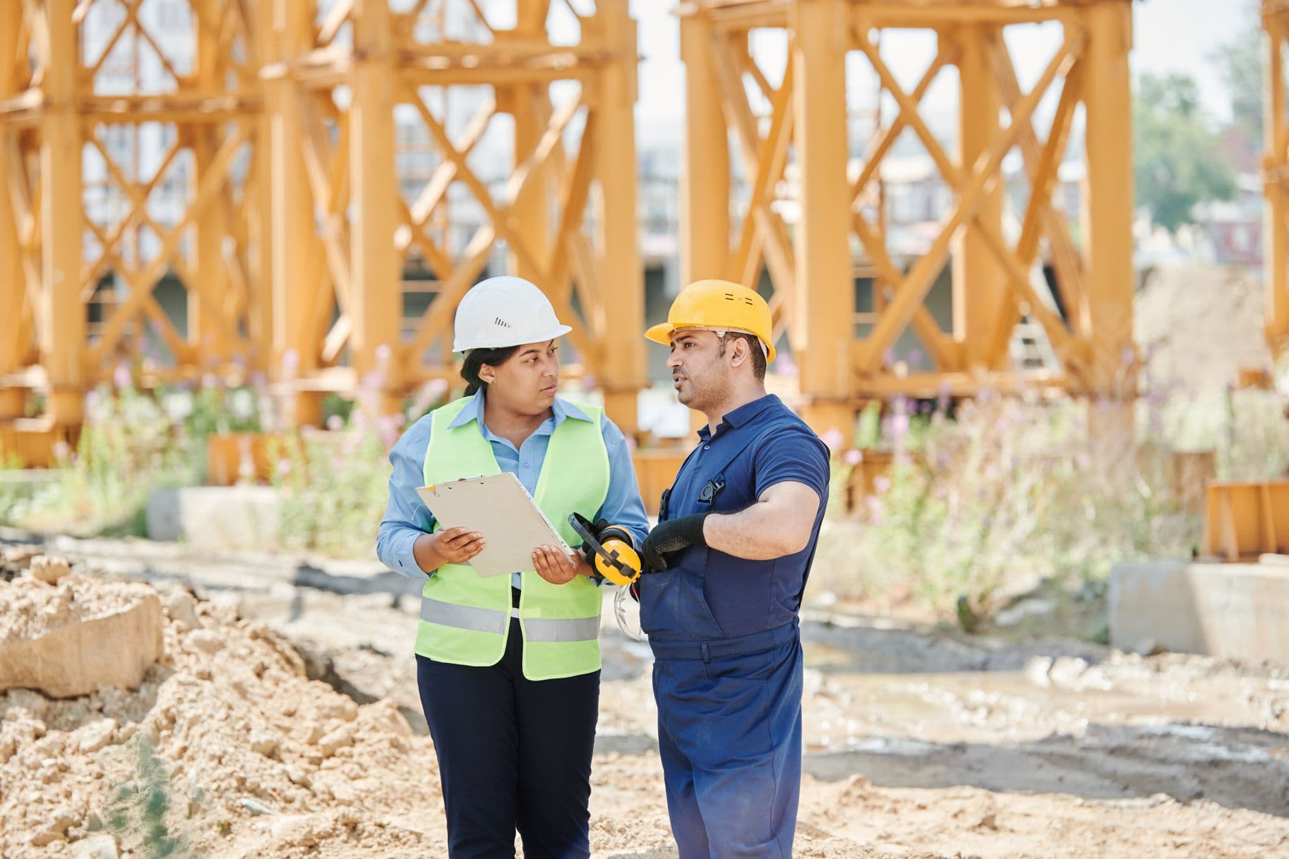 A woman in a white hard hat and hi-vis vest with a clipboard briefs a worker in a yellow hard hat at a construction site